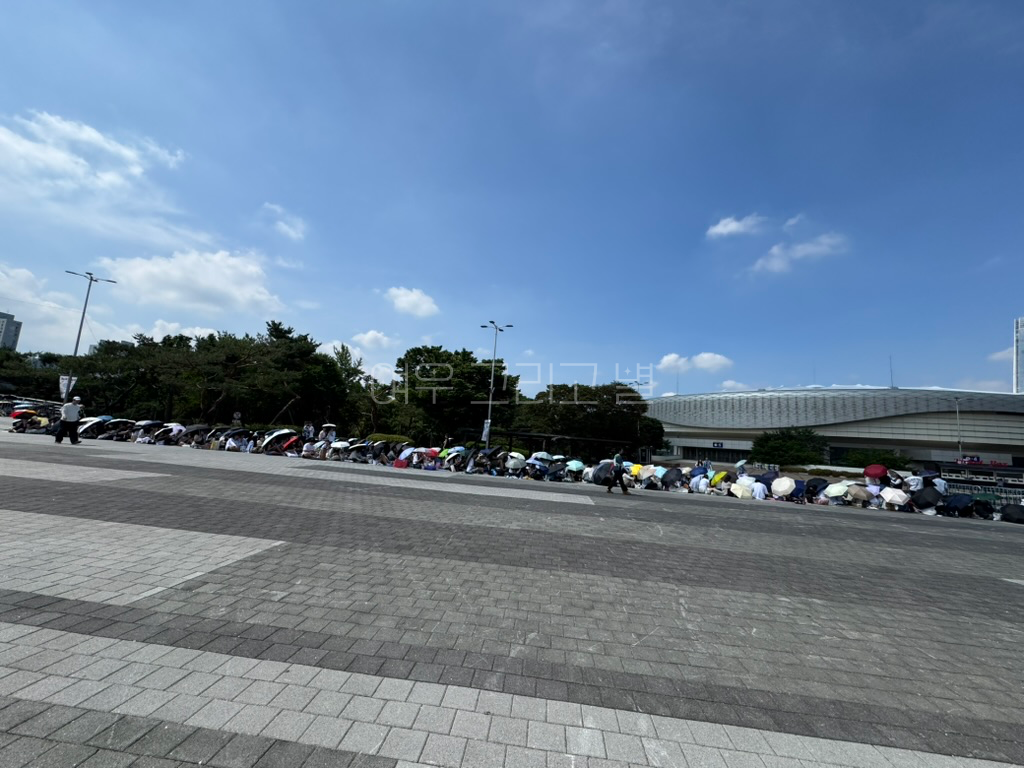 올림픽 공원에서 줄을 서있는 수많은 인파. 모두 양산을 쓰고 있다. 뒤로는 핸드볼경기장이 보이며 하늘은 구름이 많지 않고 파랗다.