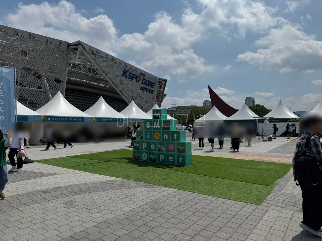 체조경기장을 배경으로 많은 부스들이 디귿자 형태로 배치되어 있다. 중간에는 인조잔디 위에 초록색으로 민트페이퍼가 적힌 정사각형 블록들이 쌓여있다.