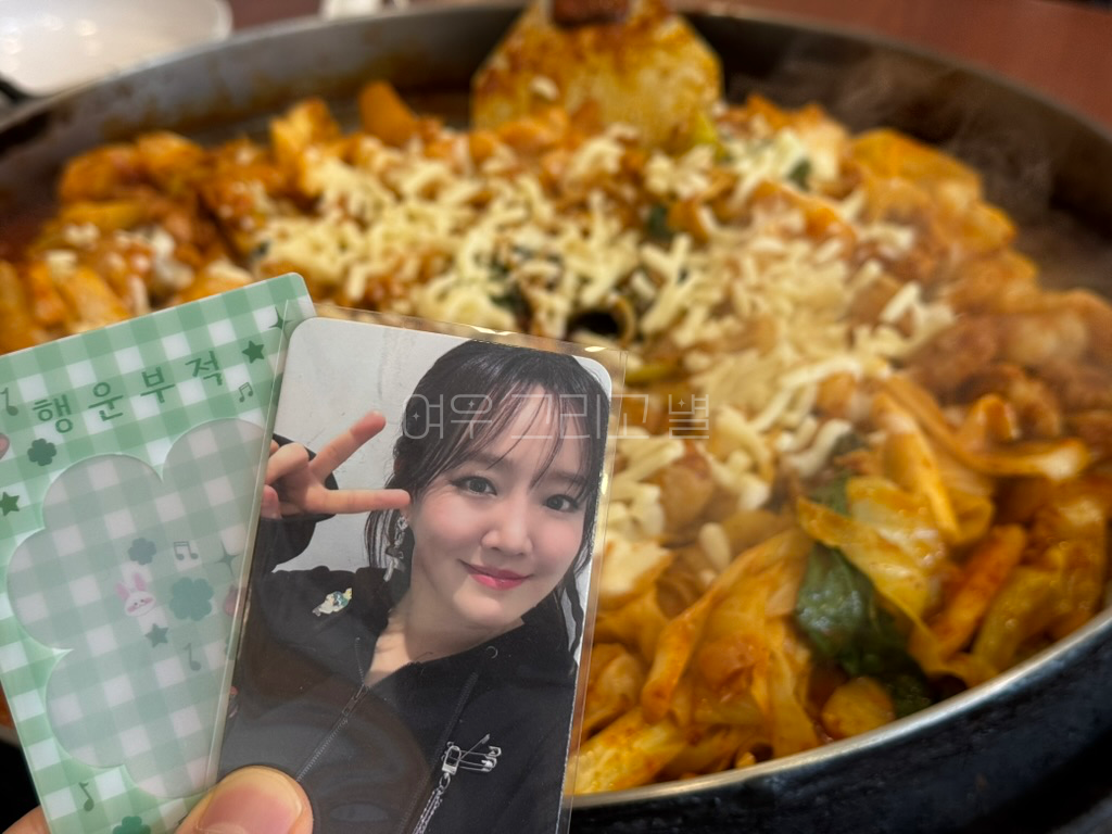 윤하교육과정평가원 2025년 생일광고 특전으로 제공된 행운부적이 적힌 초록색 체크무늬 '포토카드 L 홀더'와 동일한 이벤트 특전으로 제공된 '윤하 포토카드'를 들고 촬영한 장면이다. 뒤로는 치즈와 쫄면을 추가한 소양강 닭갈비가 보인다.
