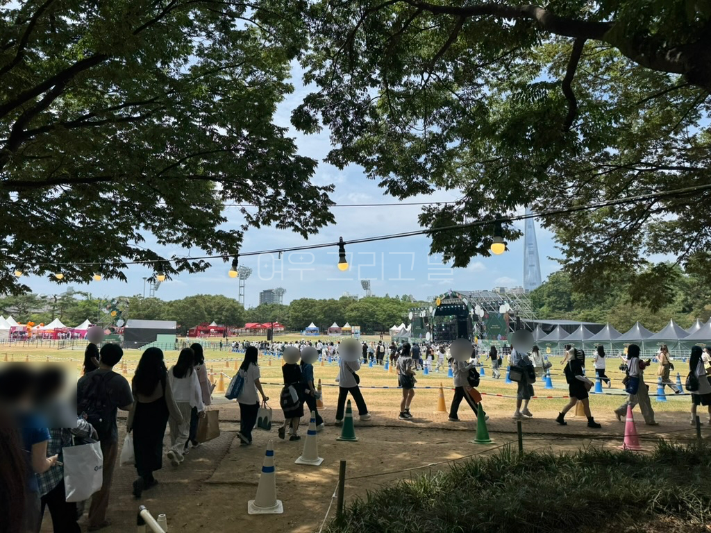 88잔디마당에 입장하고 있는 줄이 길게 이어져있다. 가는 길이 잘 정비되어 있고 나무 그늘이 형성되어 있다. 하지만 너머에 보이는 잔디마당에는 그늘 하나 찾아볼 수 없다. 배경으로는 무대와 여러가지 부스, 롯데월드타워가 보인다.